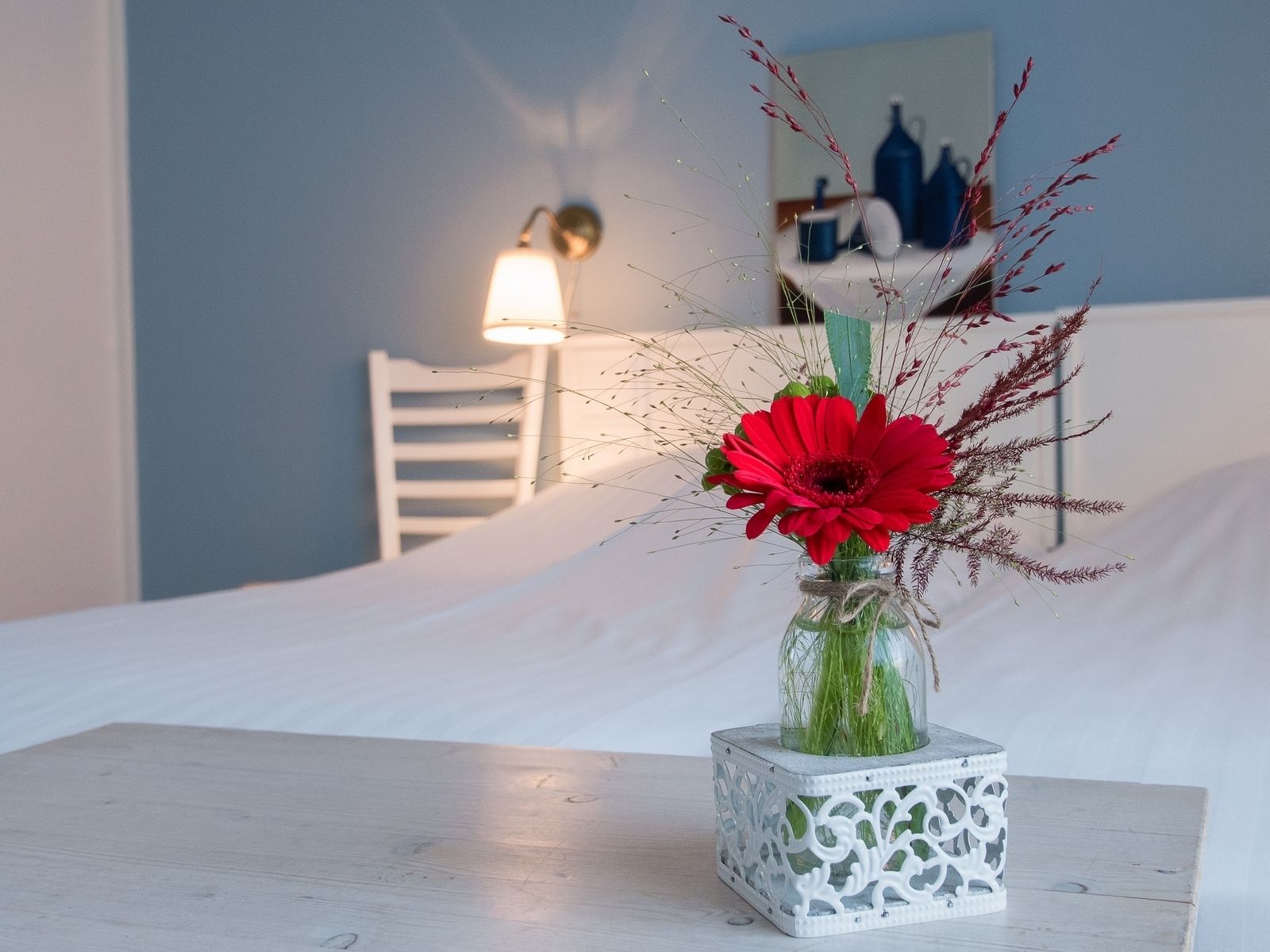 A white bed with a small white table beside it. A glass vase with red flowers and green foliage is placed on the table.