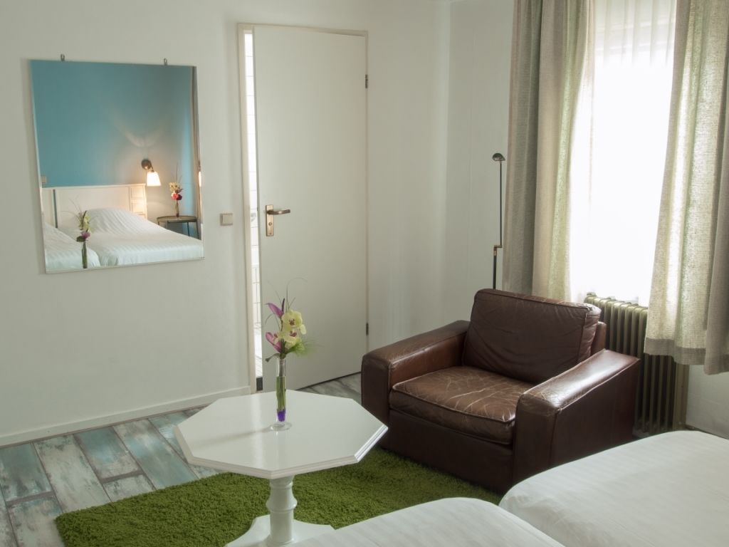 A hotel room with a brown leather sofa, a white bed, a white table with a vase, a mirror on the wall, and a window with curtains.