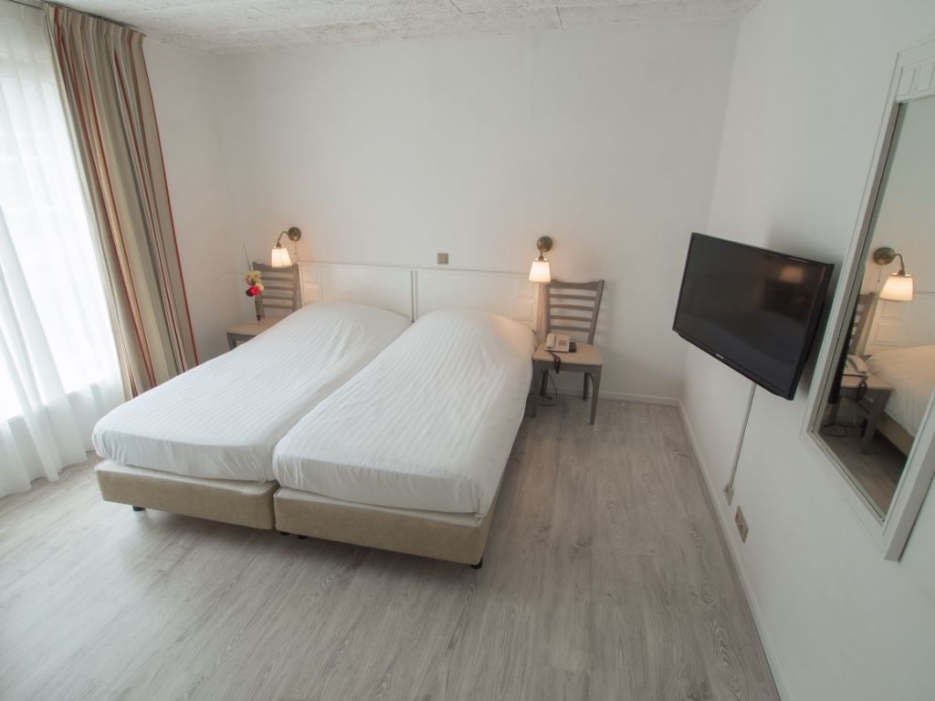 Una cama doble con estructura de madera, dos mesitas de noche con lámparas, un televisor de pantalla plana montado en la pared y una ventana con cortinas.