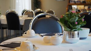 A dining table set with white teacups, saucers, and a silver teapot. There is a potted plant with pink flowers on the table.