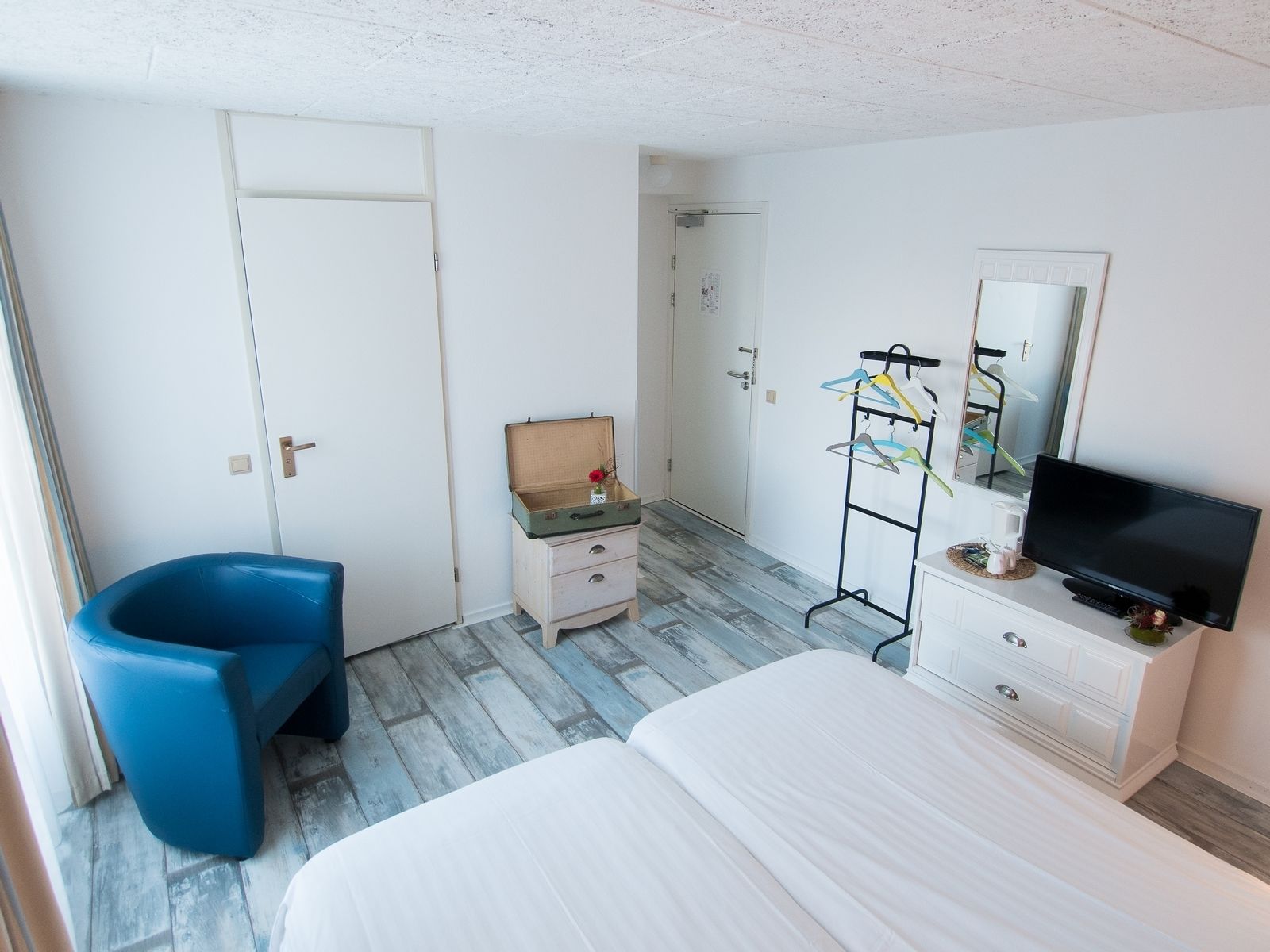 A hotel room with a bed, a small desk with a mirror, a blue chair, a coat rack, and a television.