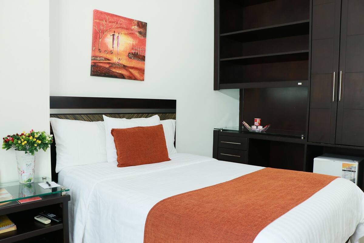 A neatly made bed with white and brown bedding, a glass side table with a vase of flowers, and a painting on the wall.