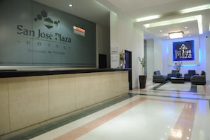 Uma área de recepção com balcão, espaço de estar com sofás e um sinal na parede para o San José Plaza Hotel.