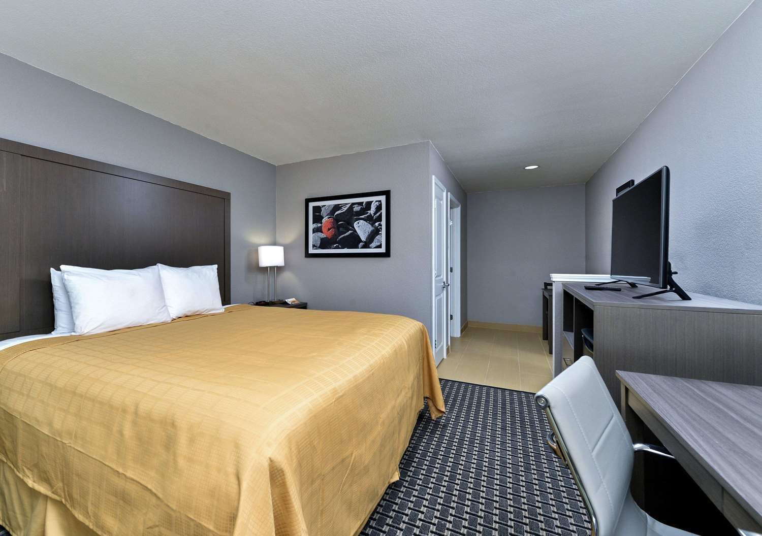 Une chambre d’hôtel comprenant un lit, une table de chevet, une lampe, une télévision, un bureau, une chaise et une porte donnant sur une autre pièce.