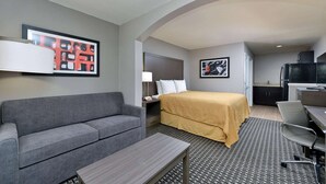 Ein Hotelzimmer mit grauem Sofa, einem Bett mit gelber Tagesdecke, einem Schreibtisch mit Stuhl, einem kleinen Tisch, einem Fernseher und Kunstwerken an den Wänden.