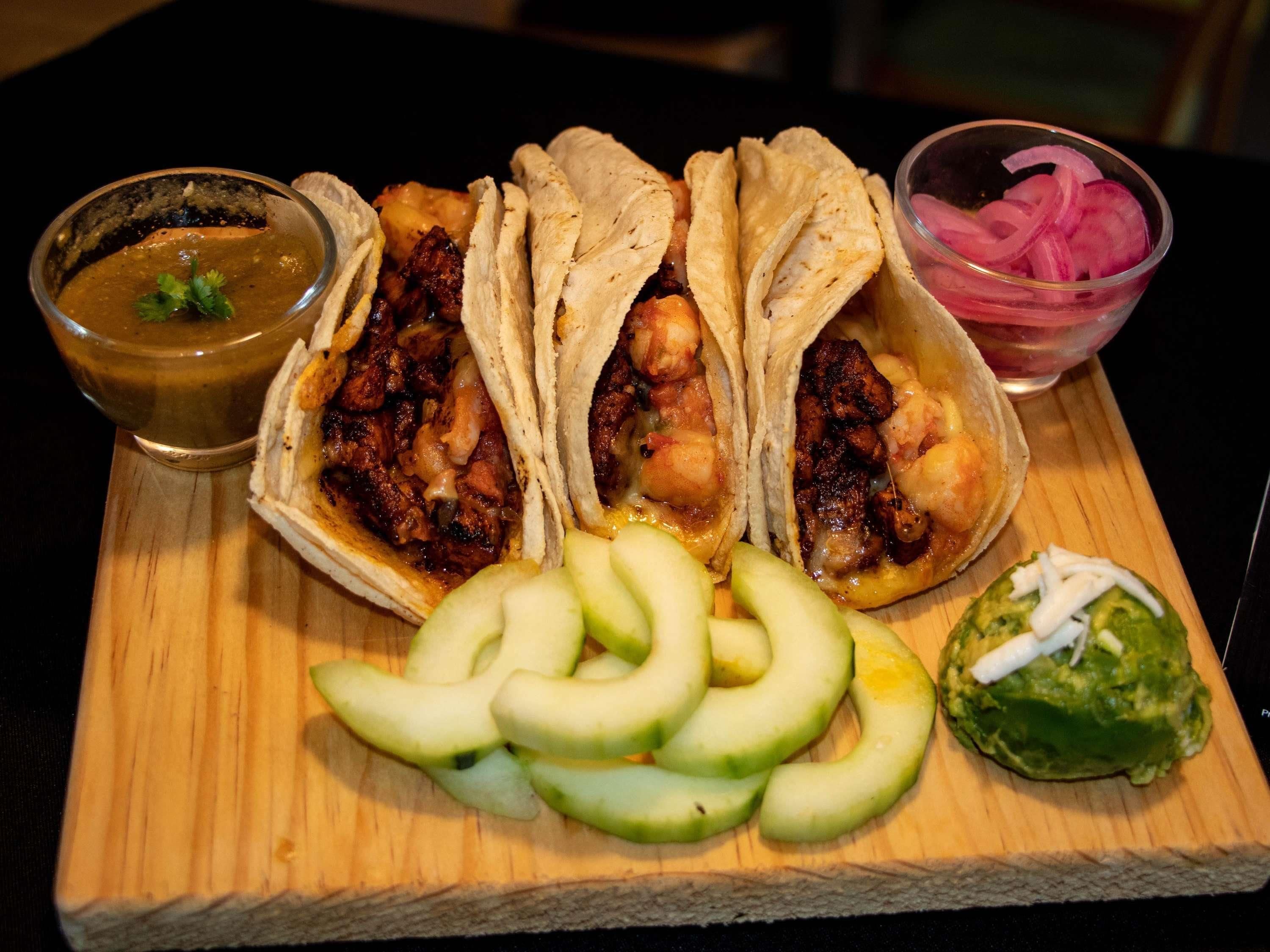 A wooden board with three tacos, a bowl of salsa, sliced avocado, and a bowl of pickled onions.