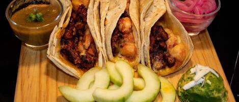A wooden board with three tacos, a bowl of salsa, sliced avocado, and a bowl of pickled onions.