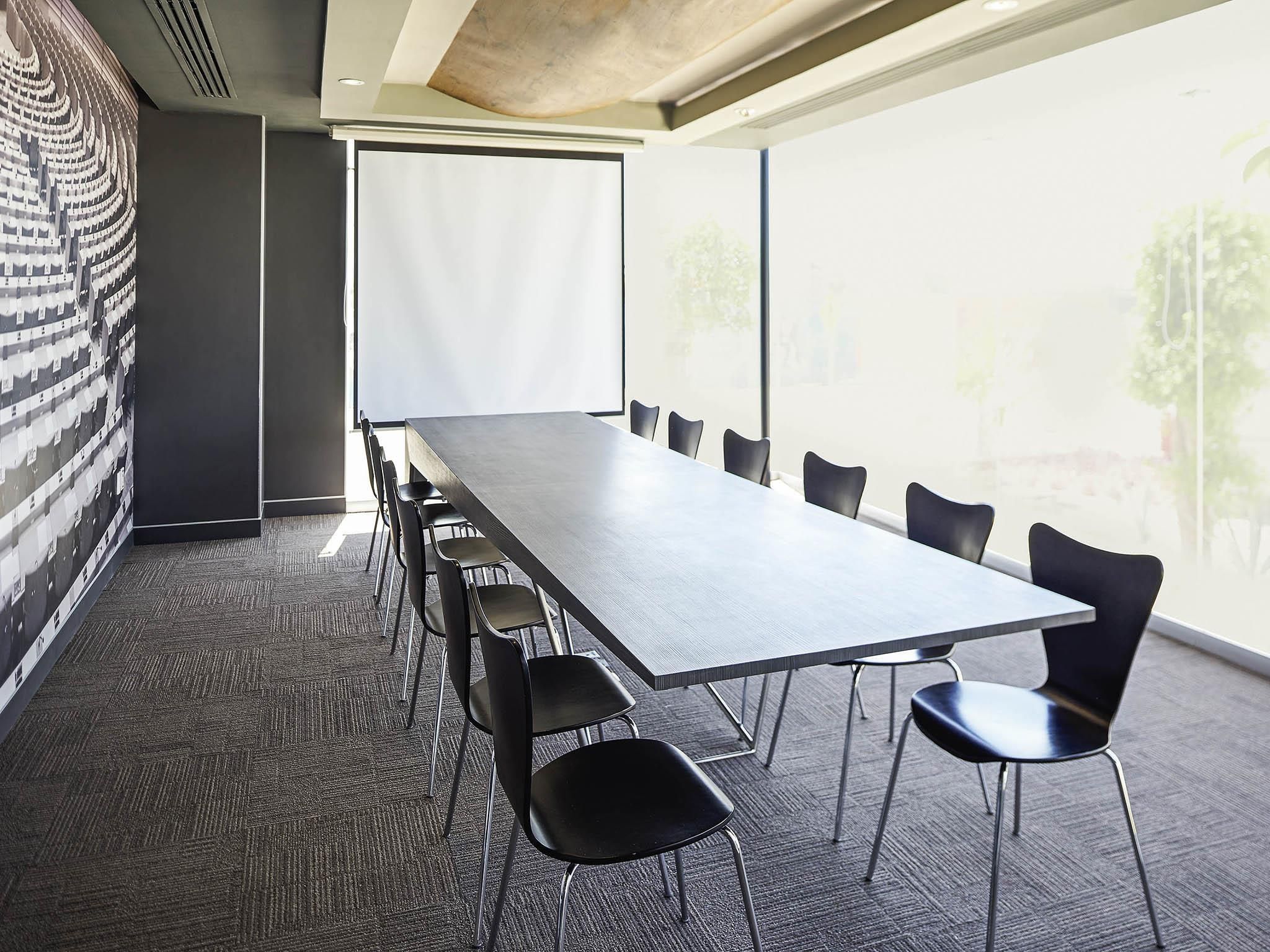 Un salón de conferencias con una larga mesa y sillas, una pantalla de proyector y ventanas amplias.