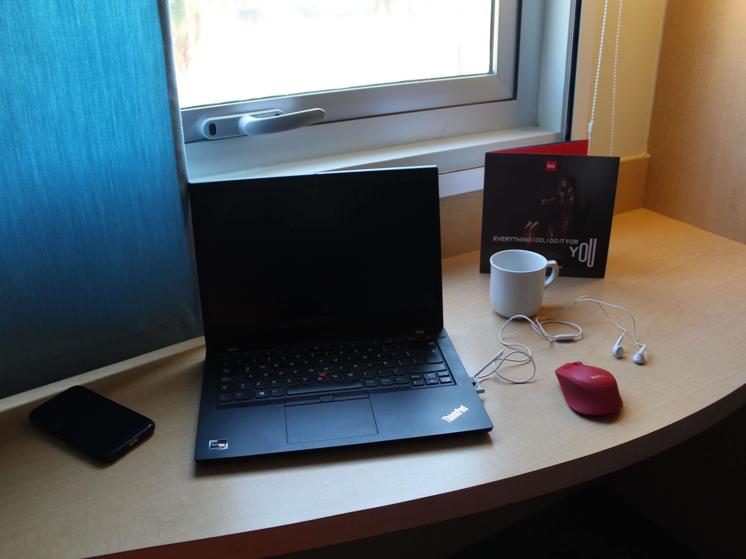 Um laptop sobre a mesa perto de uma janela, um smartphone, uma caneca branca, um par de fones de ouvido e um mouse vermelho.