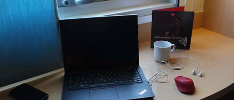 Um laptop sobre a mesa perto de uma janela, um smartphone, uma caneca branca, um par de fones de ouvido e um mouse vermelho.