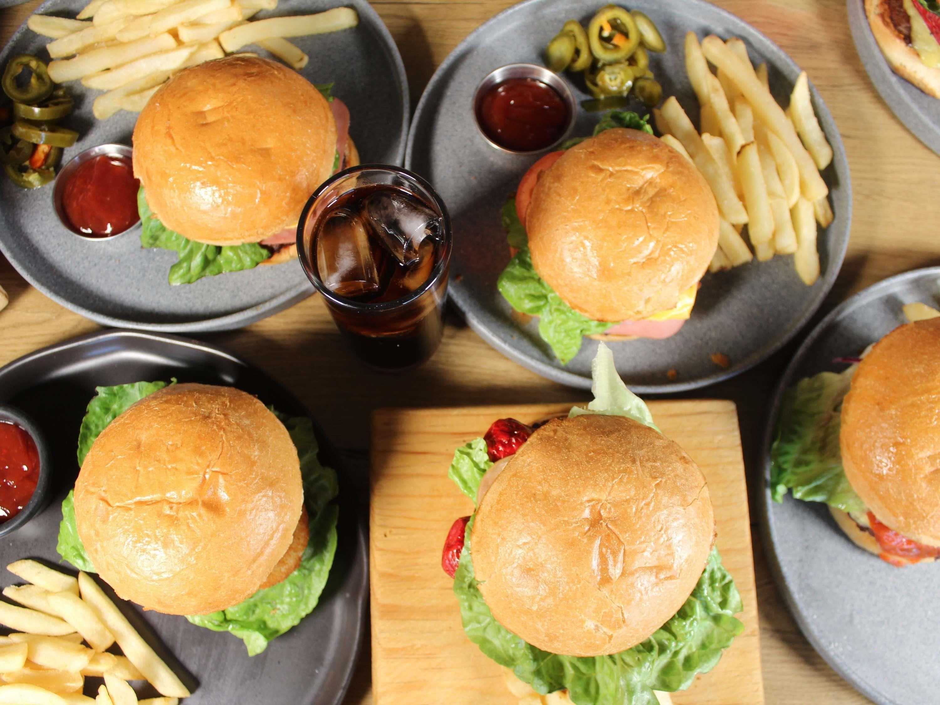 Una mesa con cuatro hamburguesas, cada una acompañada de papas fritas y salsas, y un vaso de refresco.