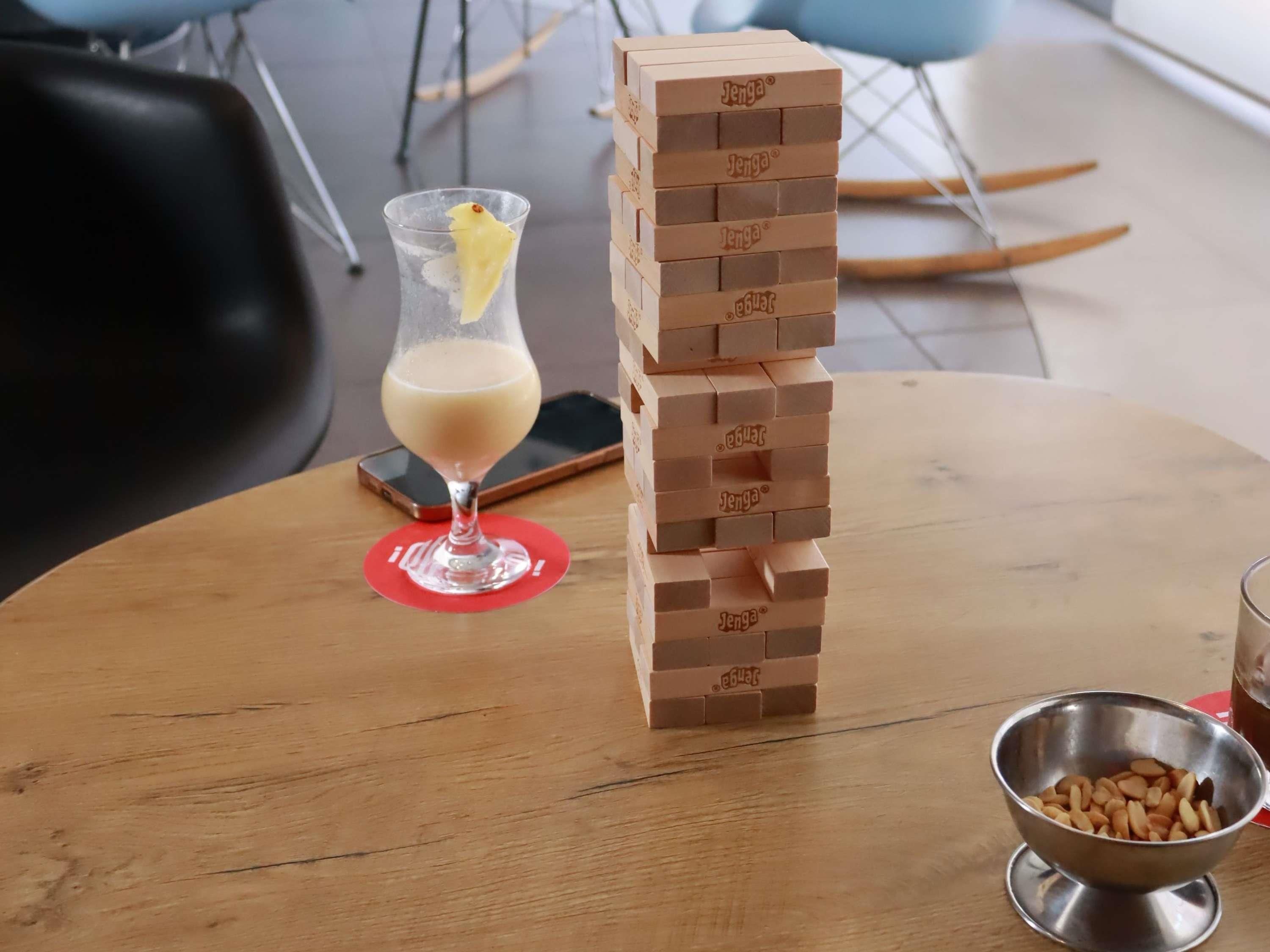 Une table en bois sur laquelle se trouve une tour de Jenga, un verre contenant une boisson crémeuse agrémentée d’une tranche de citron, et un bol en métal rempli de noix.