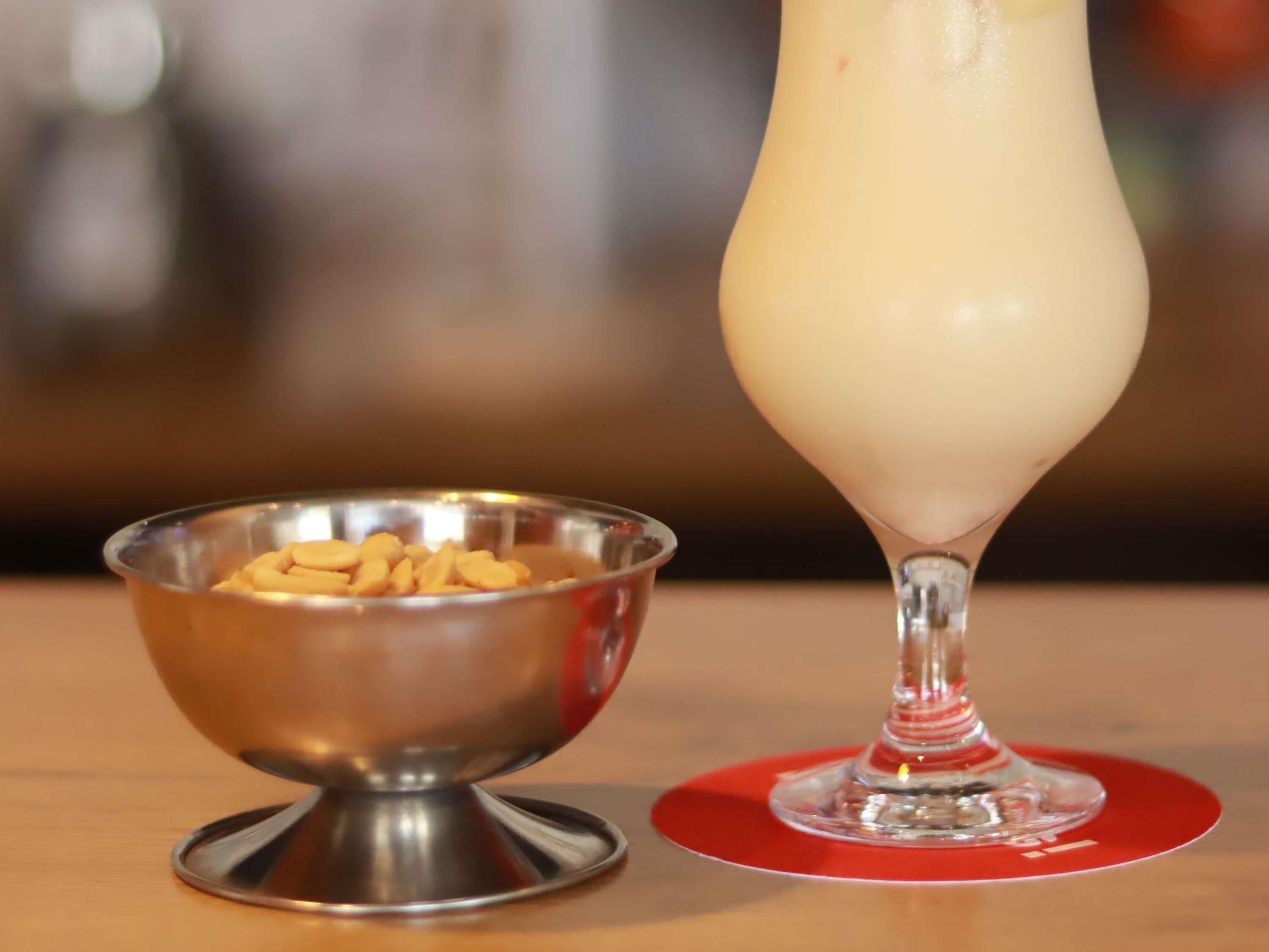 A clear glass of a creamy beverage next to a metal bowl with nuts on a wooden table.