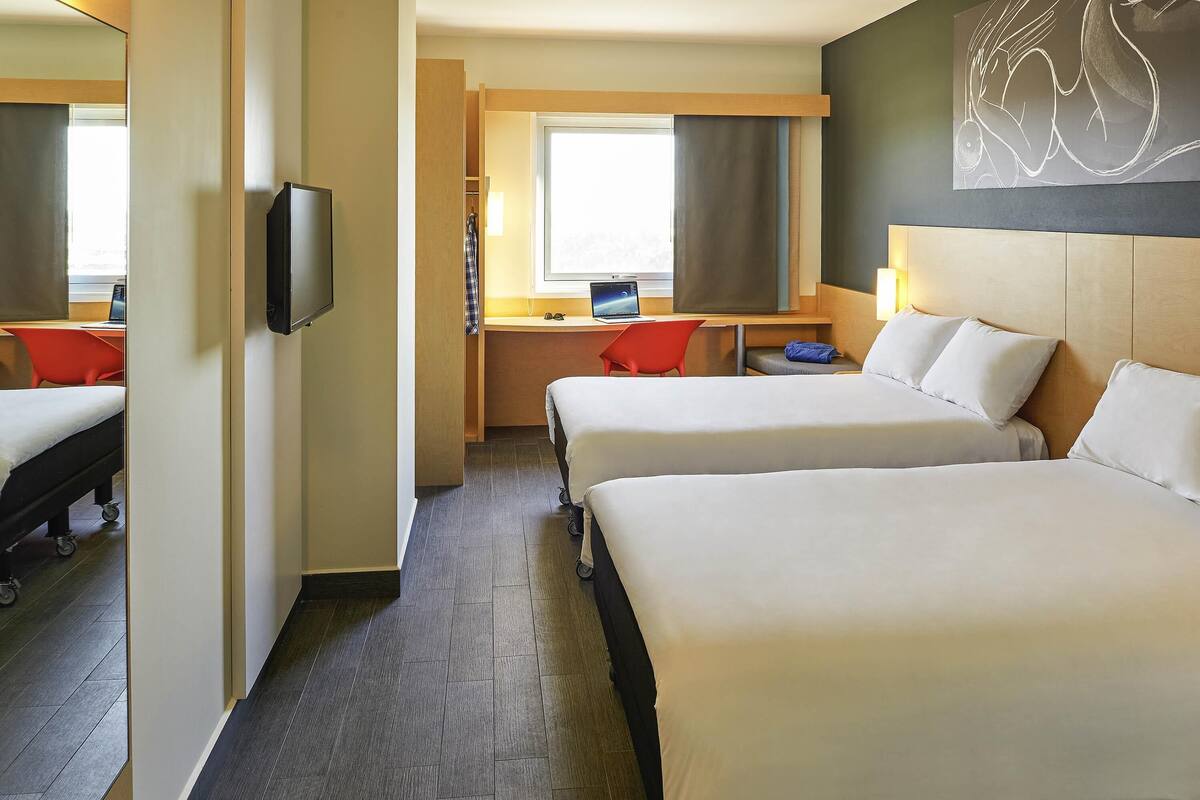 Una habitación de hotel con dos camas, un escritorio con computadora, un televisor y un ventanal grande.
