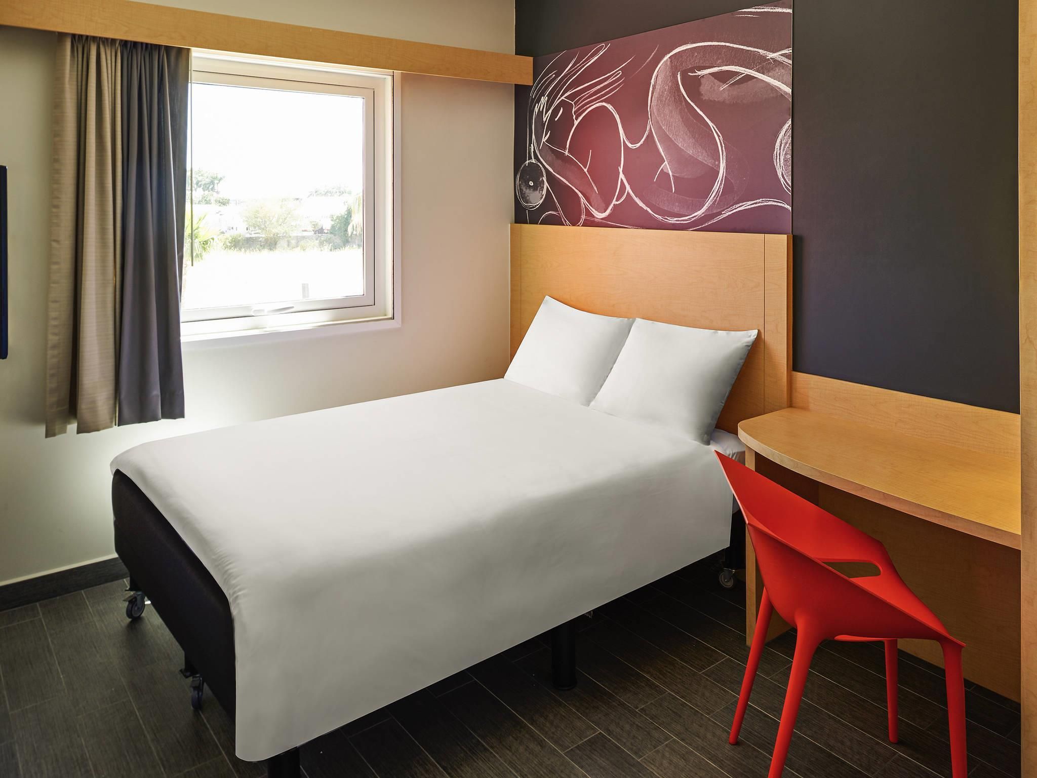 Une chambre d’hôtel comprenant un lit, un bureau, une chaise, une fenêtre avec des rideaux et un mur orné d’un motif décoratif.