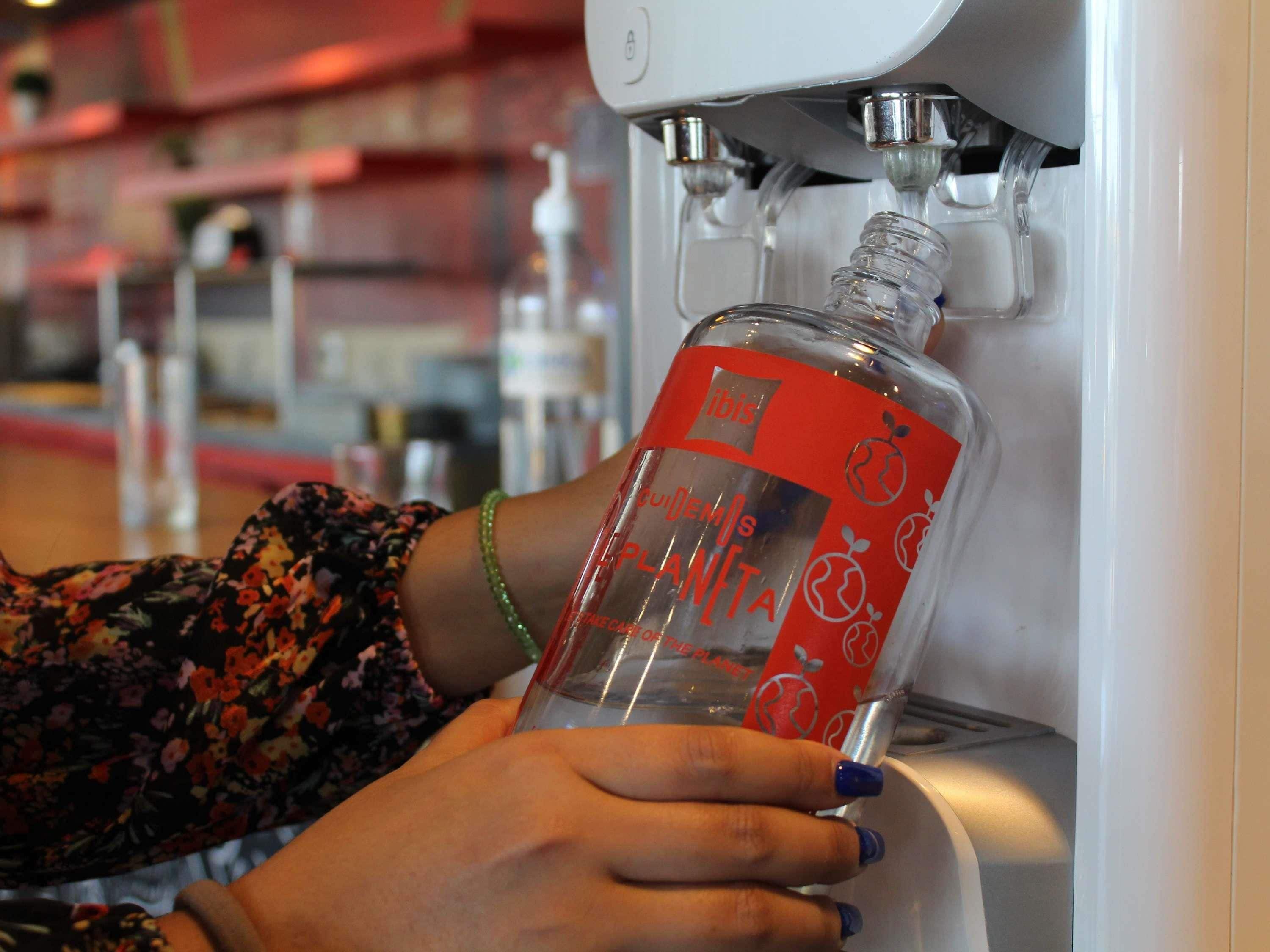 Una mano sostiene una botella con etiqueta roja, ubicada debajo de un dispensador de agua.