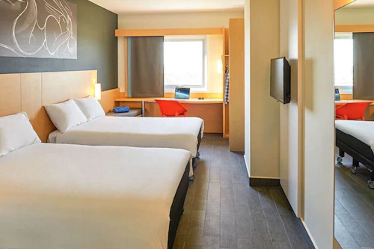 A hotel room with two beds, a desk, a chair, a television, and a large window.
