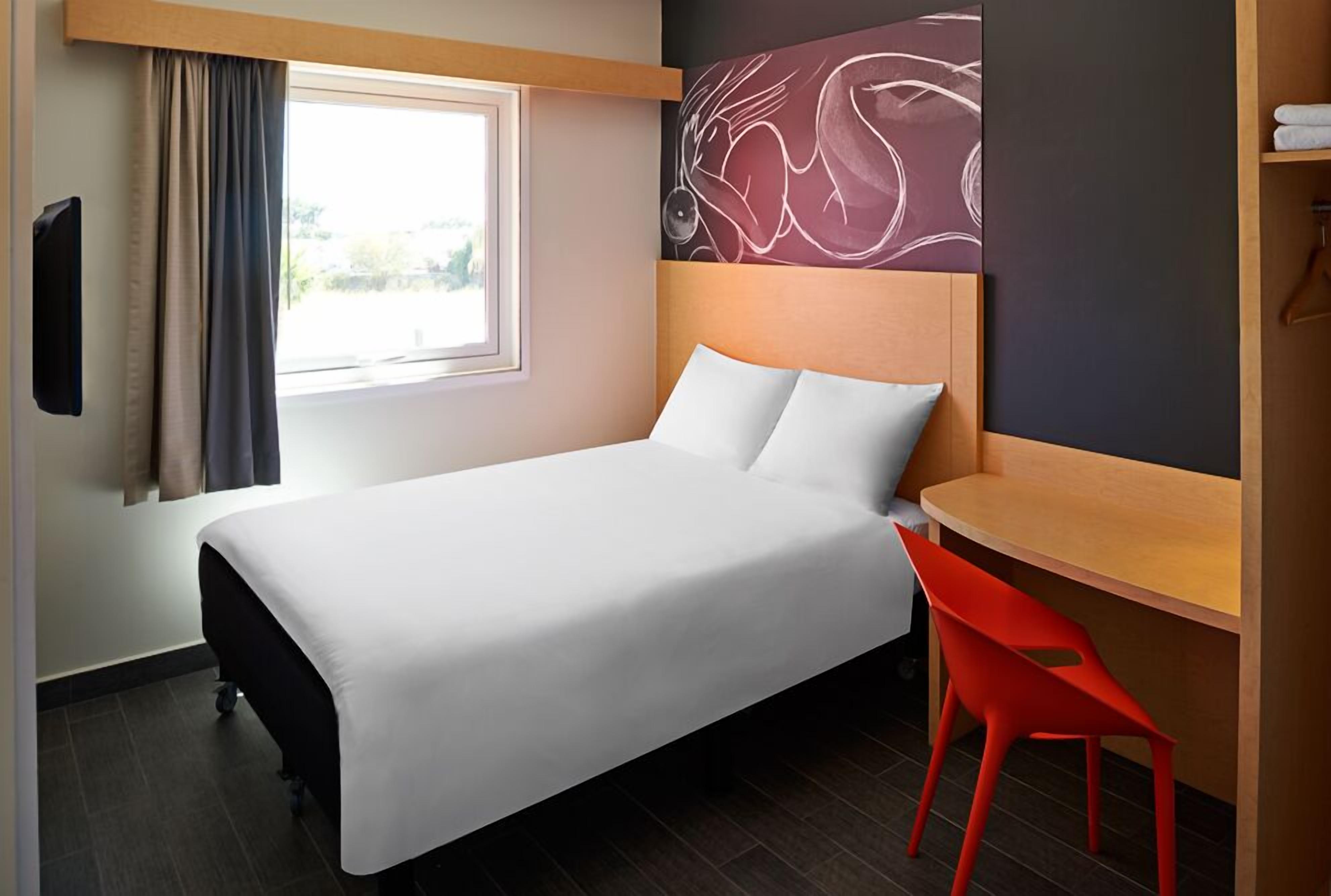 Una habitación de hotel con cama, escritorio, silla, televisor y ventana con cortinas.