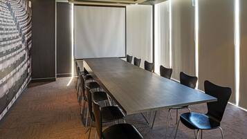 Un salón de conferencias con una larga mesa y sillas, ventanas amplias y un muro decorado.