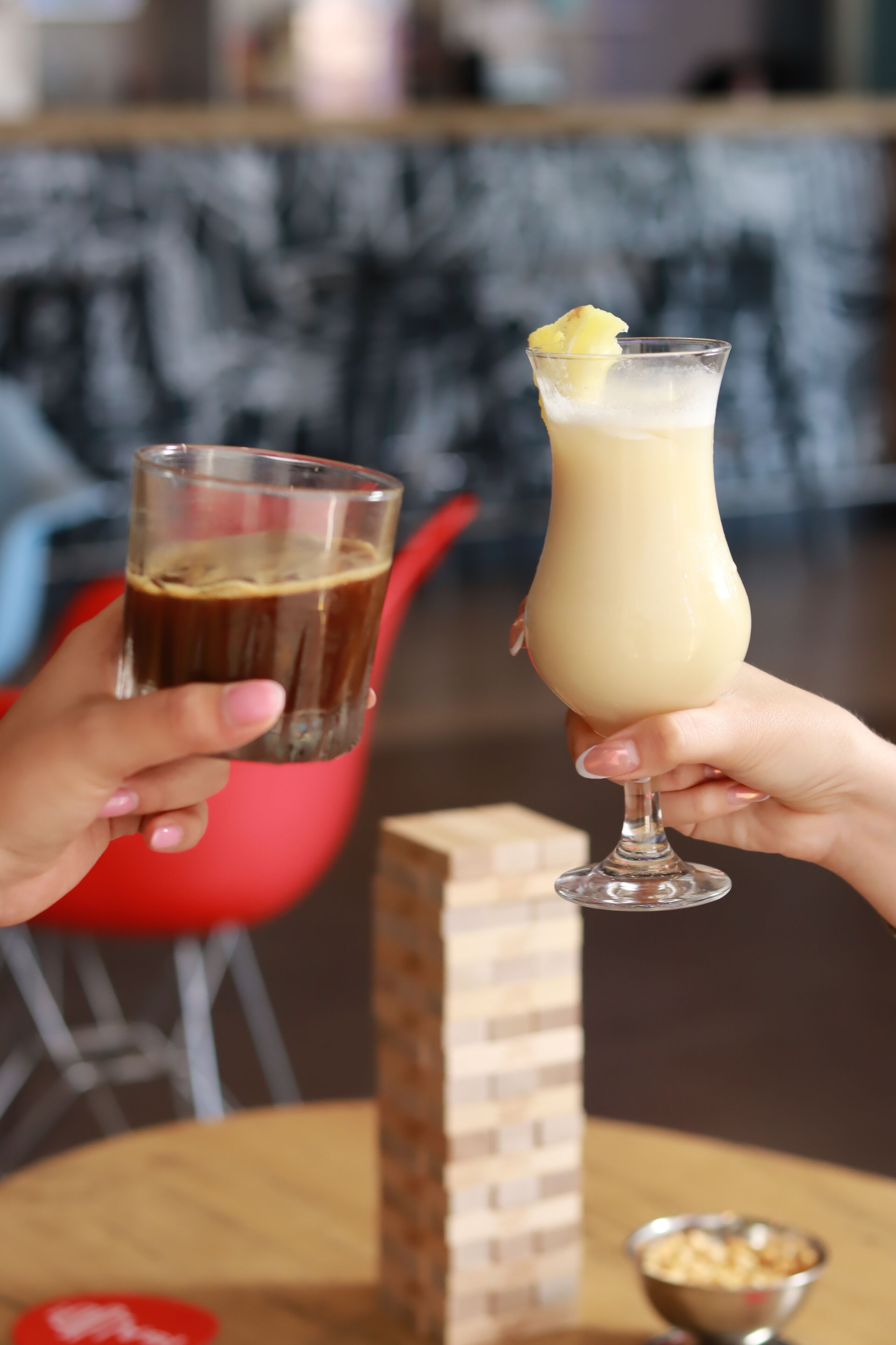 Two hands holding glasses of a beverage, a red chair, and a small bowl of popcorn in the background.