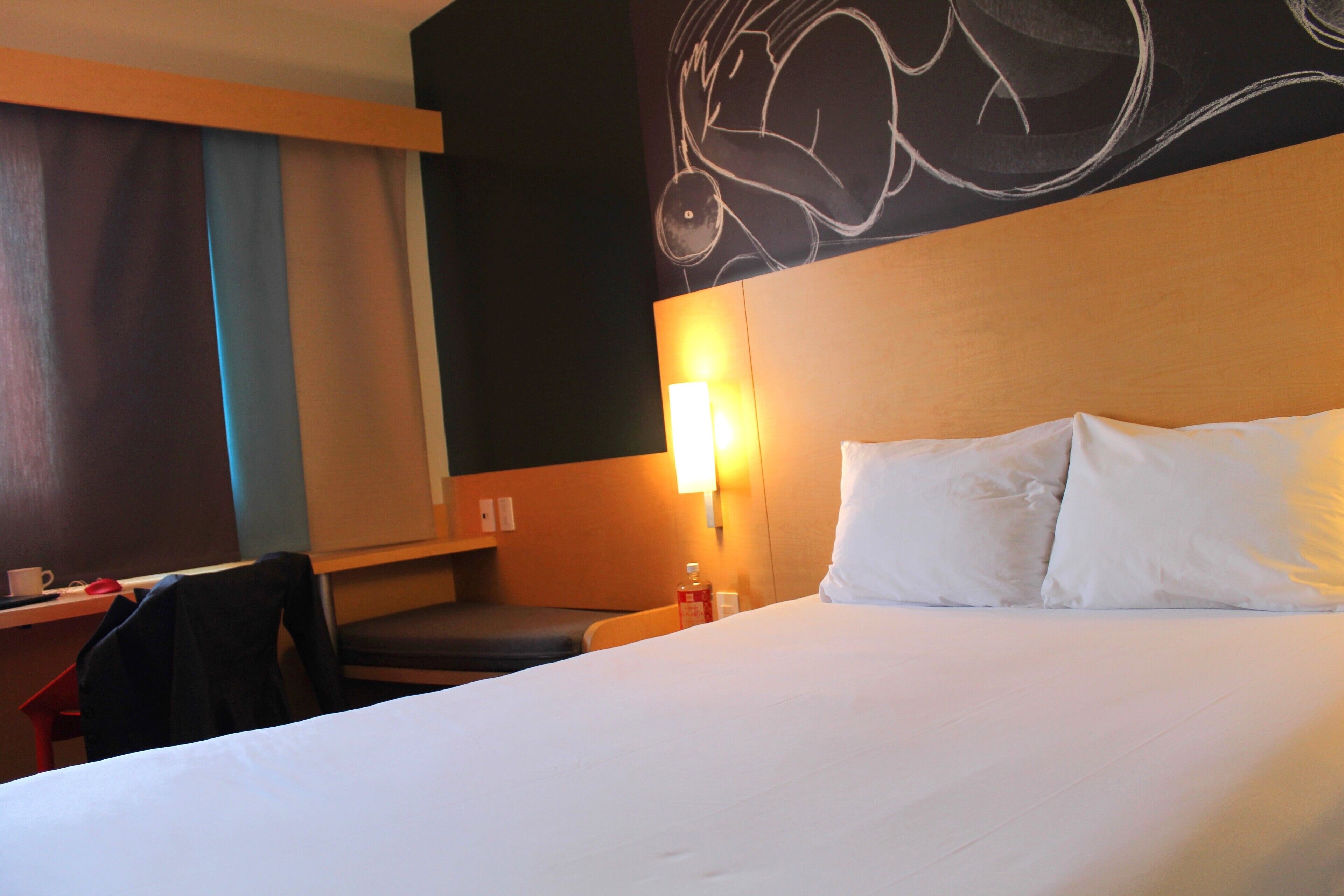 Habitación de hotel con una cama grande, un escritorio con una silla, una lámpara de pared y una vista parcial de una ventana con cortinas.