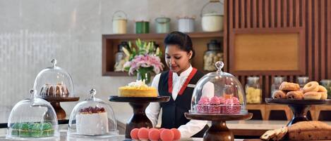 A buffet setup with various desserts under glass domes, a person in uniform, and kitchen shelves in the background.