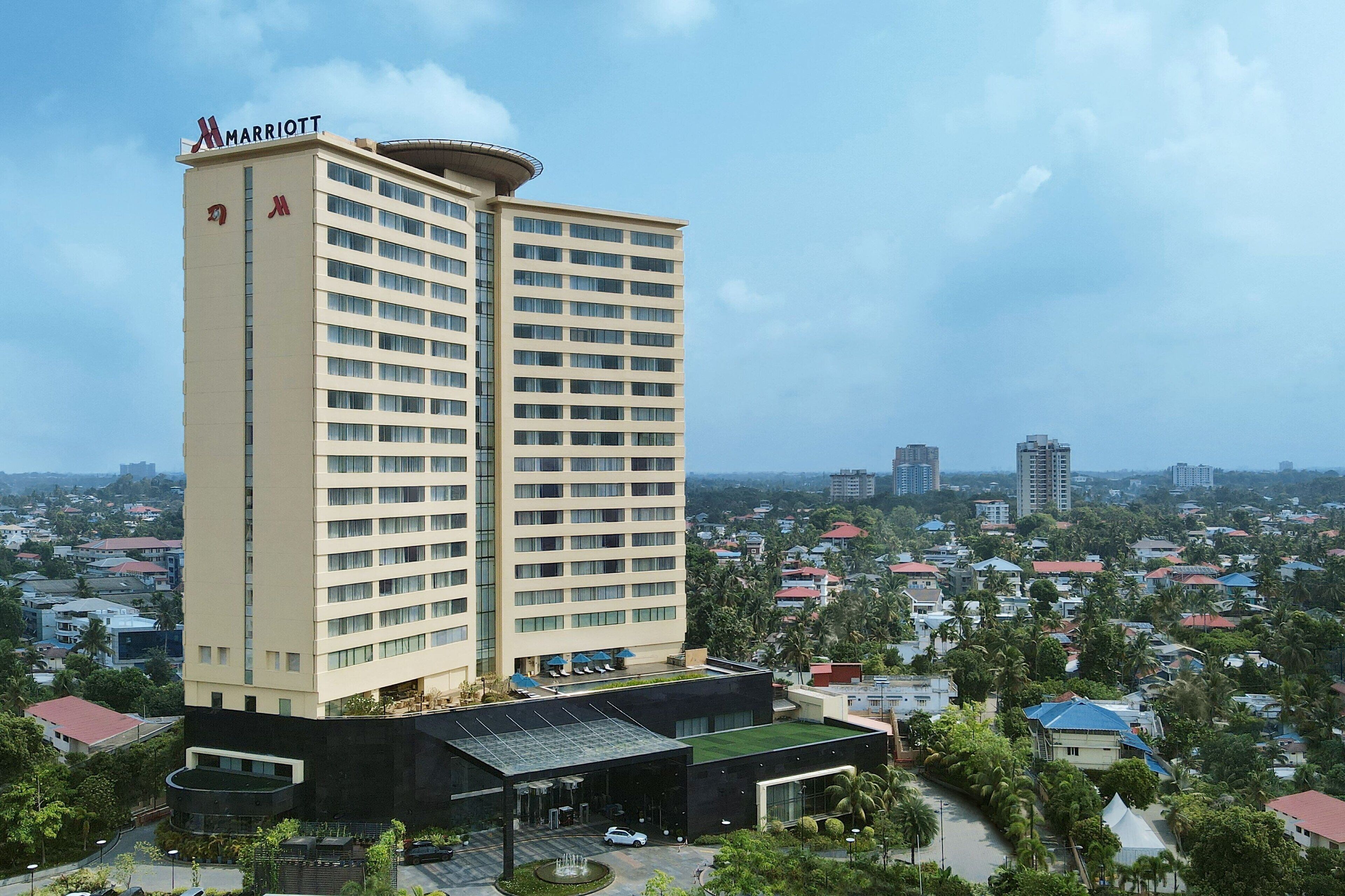 Un hotel alto de varios pisos, de color beige y diseño moderno, con un llamativo logotipo de 'Marriott', rodeado de un exuberante paisaje verde y edificios residenciales.