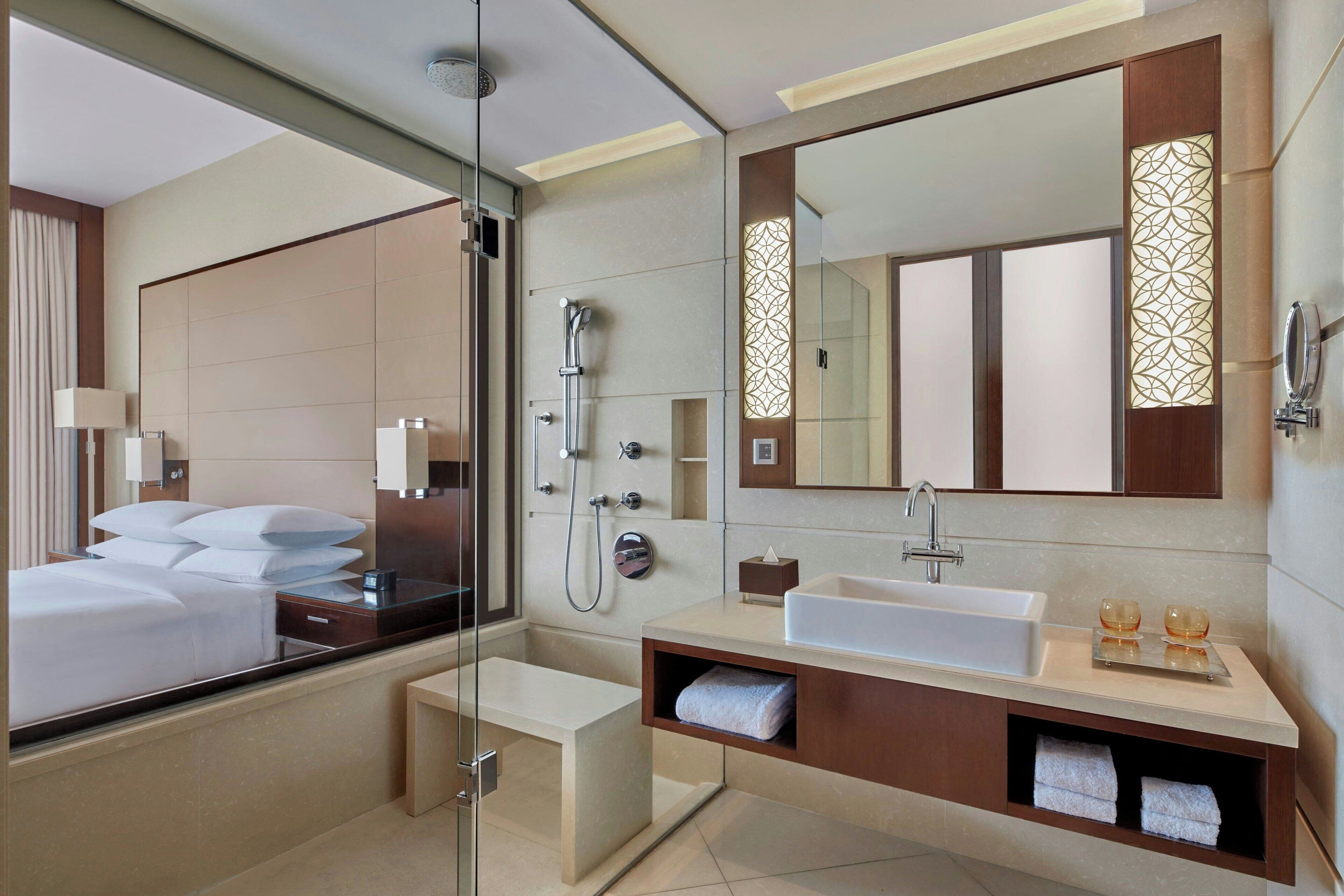 Ein modernes Hotelzimmer mit einem großen Bett, einer in Glas eingefassten Dusche, einem Schminktisch mit Waschbecken und einem großen Spiegel.