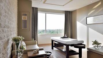 A modern spa room with a massage table, a cushioned seating area, and a large window offering a view of the outdoors.