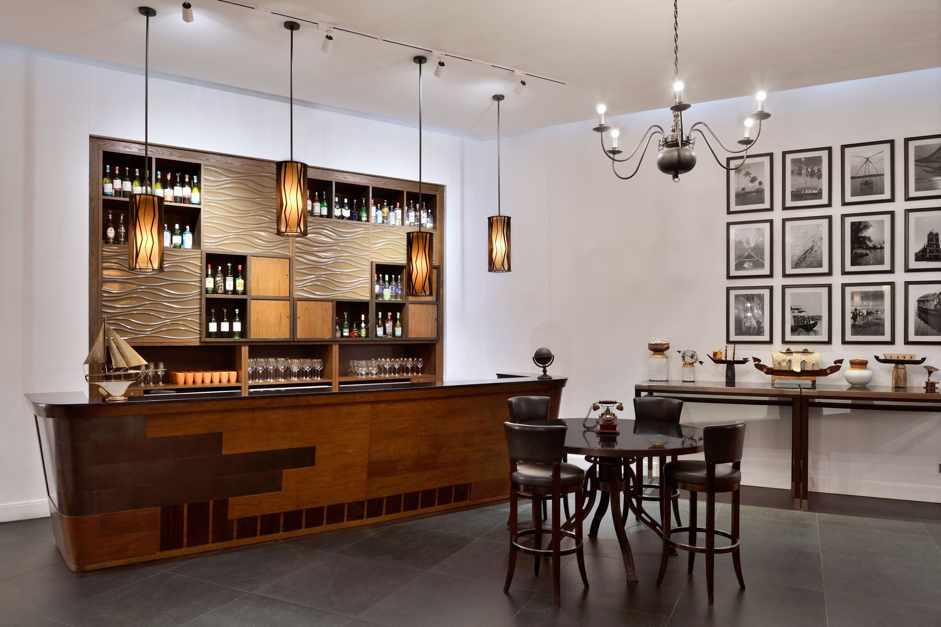 A bar area with a wooden counter, multiple shelves of bottles, and hanging pendant lights. There are chairs and a table with a globe and decorative items.