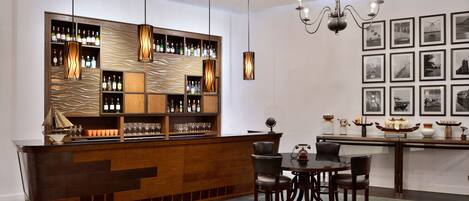 A bar area with a wooden counter, multiple shelves of bottles, and hanging pendant lights. There are chairs and a table with a globe and decorative items.