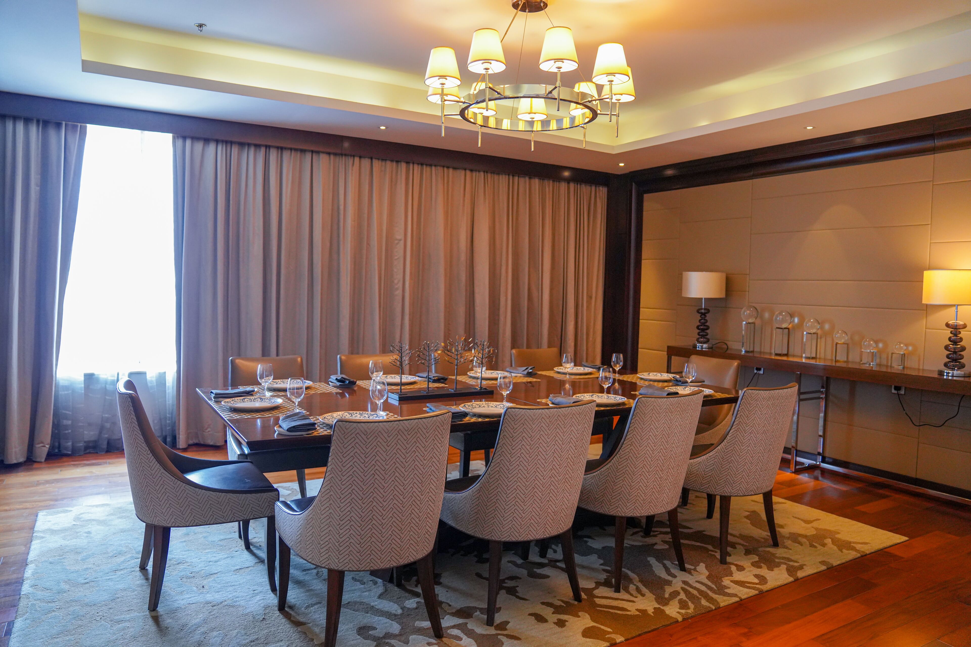 Un comedor con una larga mesa dispuesta para seis comensales, rodeada de sillas estampadas, un candelabro y vistas al exterior a través de amplios ventanales.