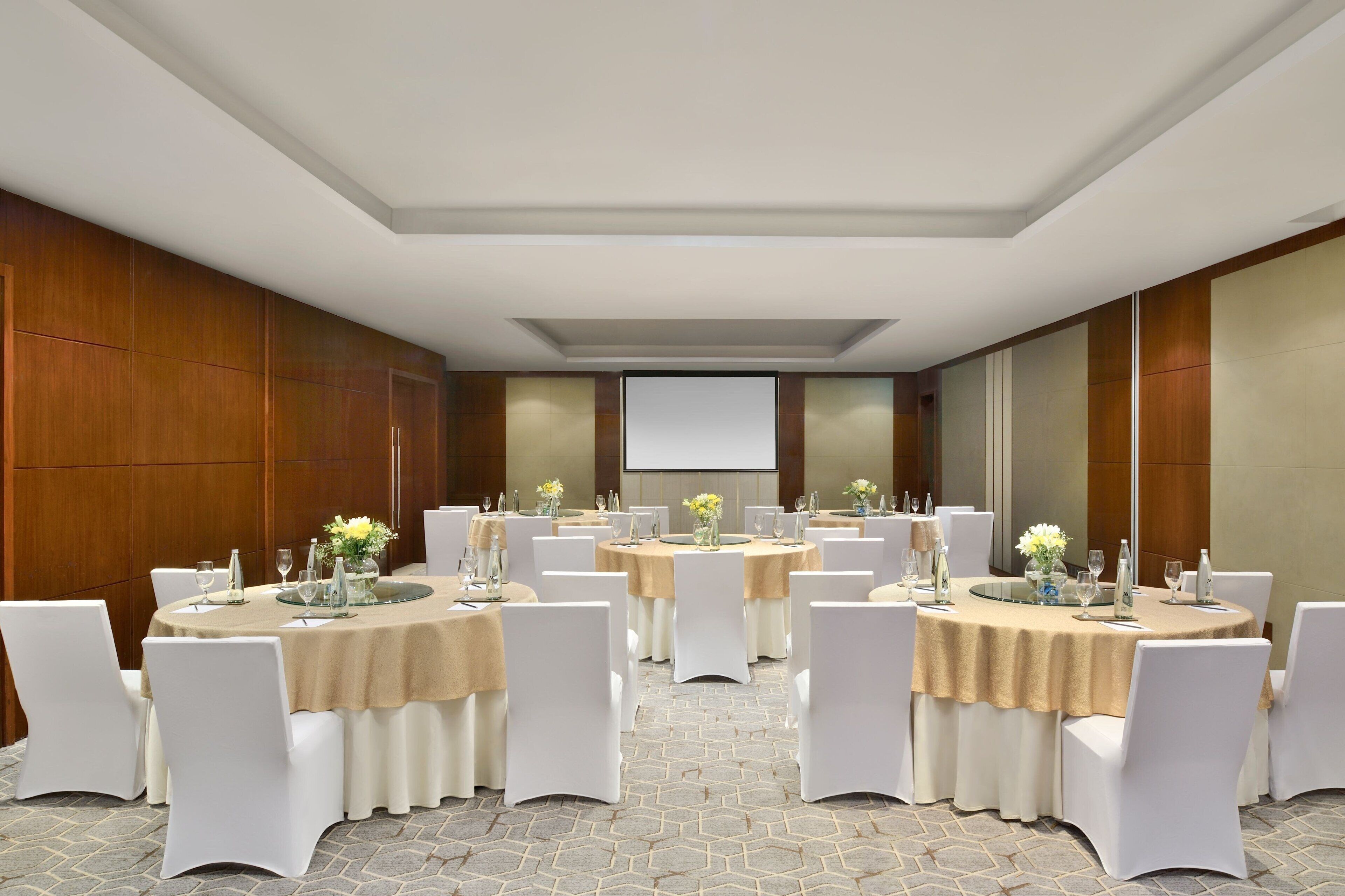A conference room with multiple round tables, each covered with a beige tablecloth and surrounded by white chairs.