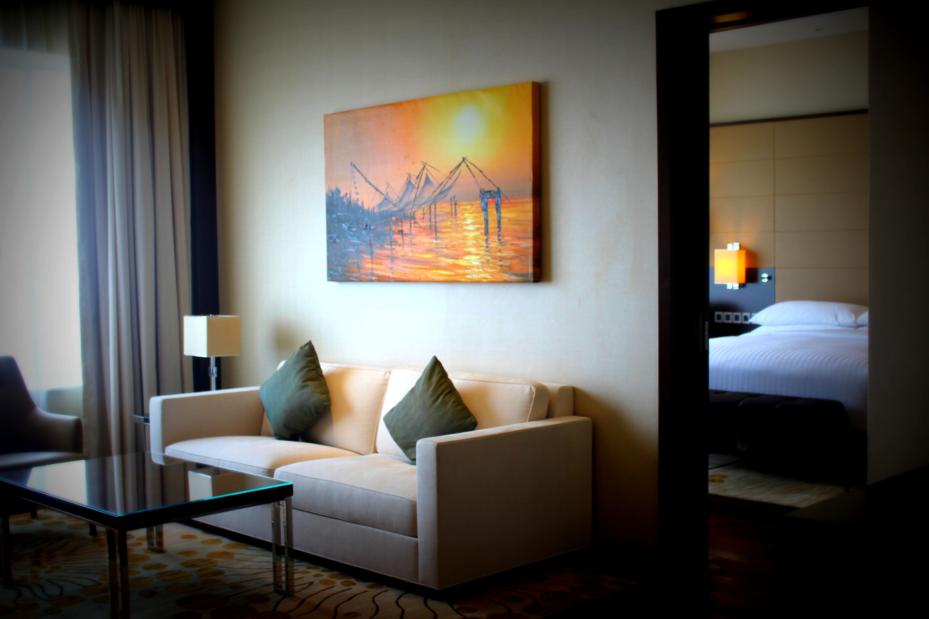 A hotel room with a sofa, a painting of a sunset, a bed, and a nightstand with a lamp.