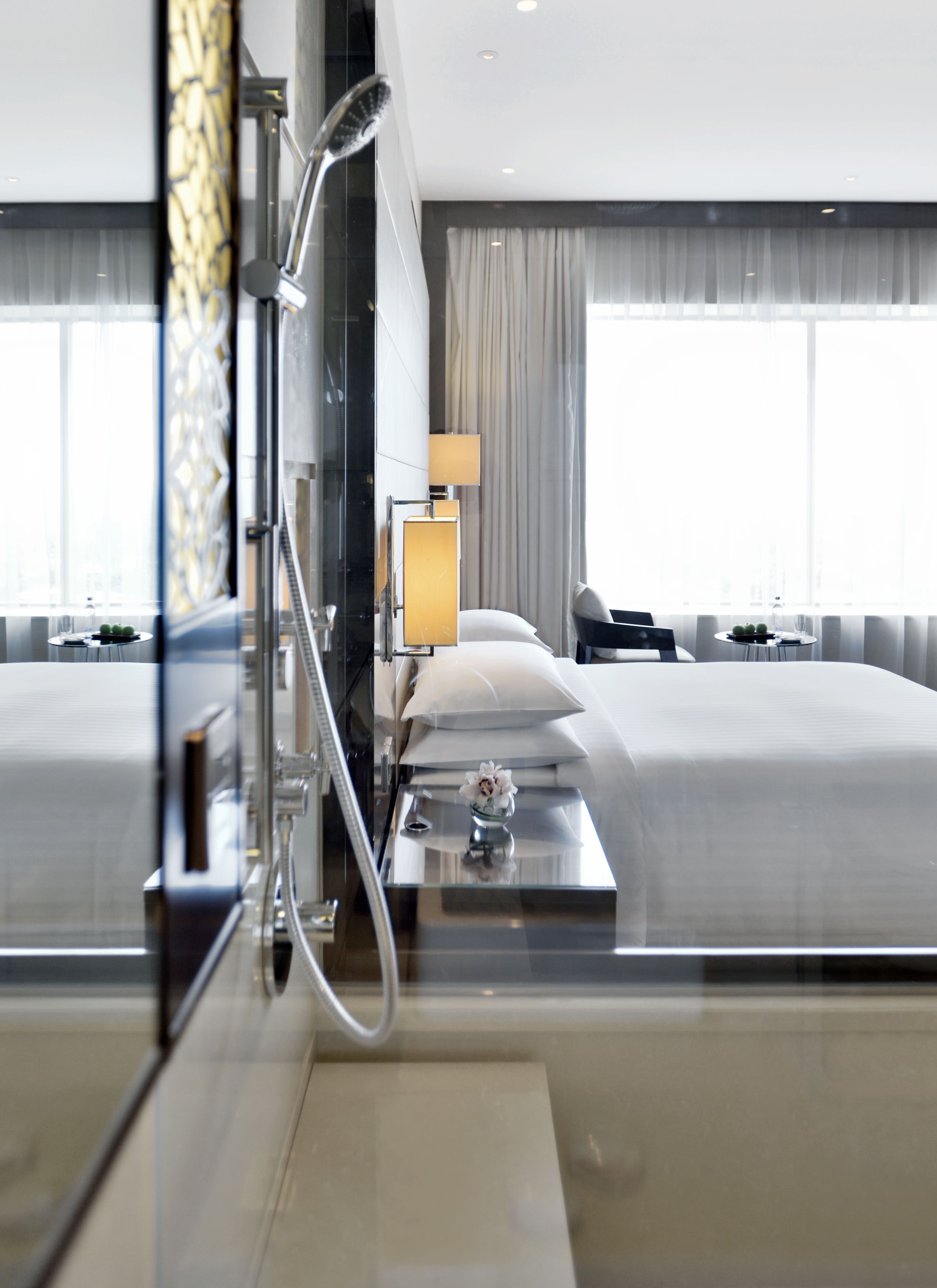 A modern hotel room with a large bed, a shower, a bedside table with a lamp, and a view of the city through large windows.