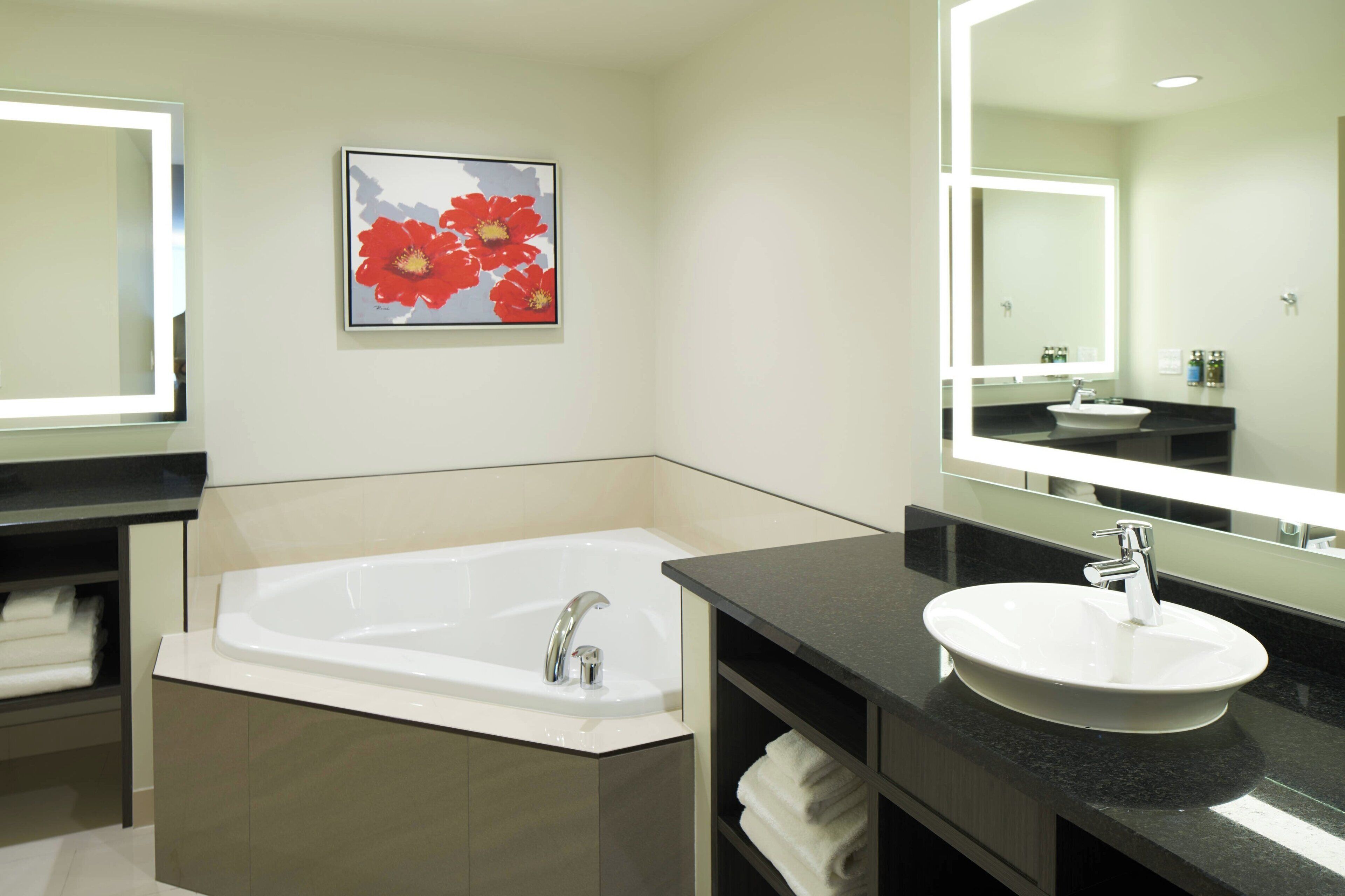 A modern bathroom with a bathtub, a sink, and a large mirror. There is a framed picture of flowers on the wall.
