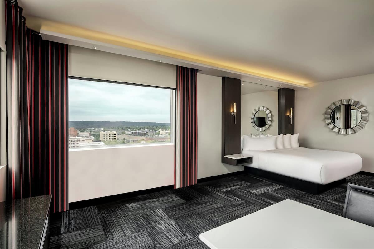 A modern hotel room with a large window, a bed, a bedside table, and two wall-mounted mirrors.