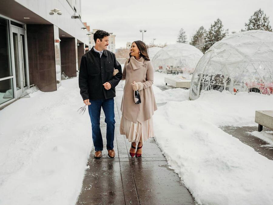A couple walking on a snow-covered path with a modern building and transparent structures in the background.