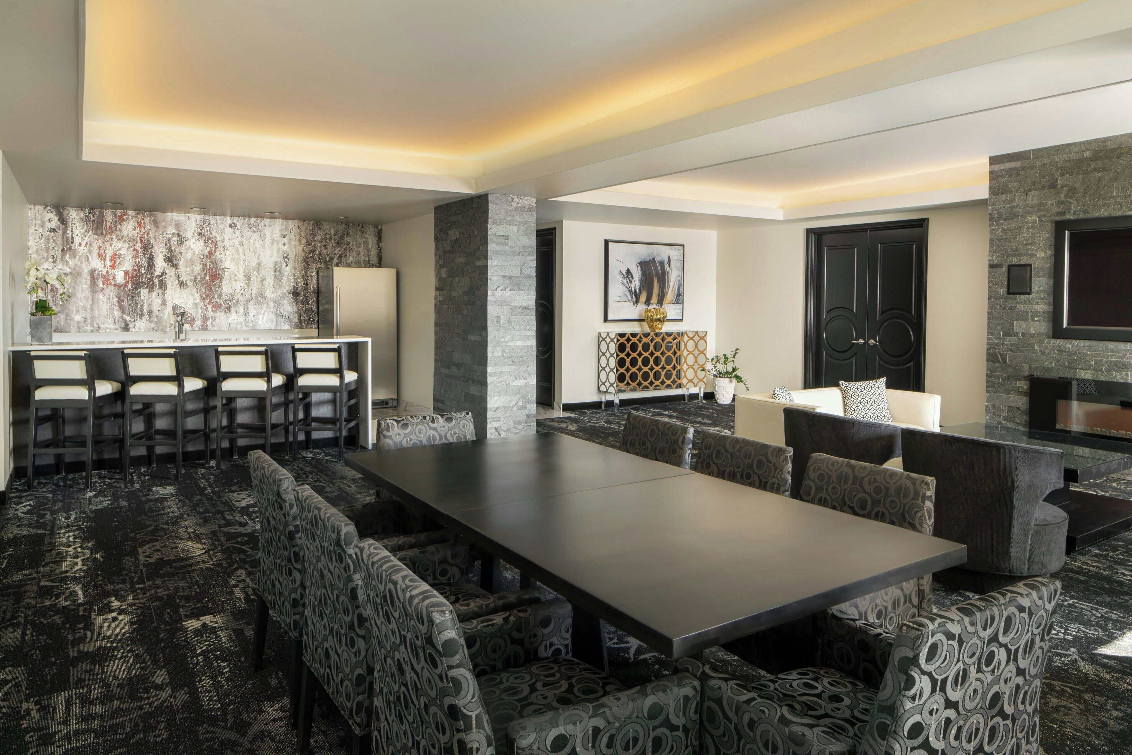 A modern dining area with a long table and patterned chairs, a kitchen with a stone backsplash, and a black-framed television on the wall.
