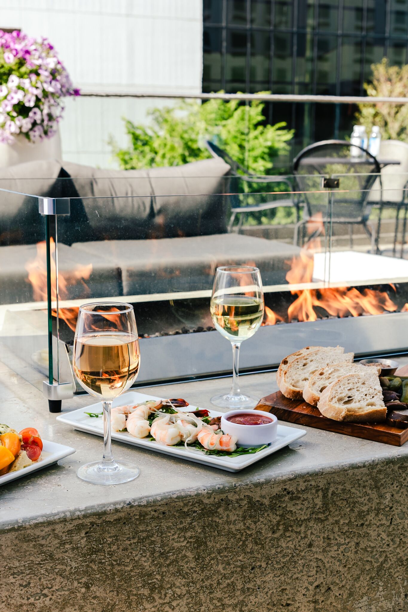 An outdoor dining area with a glass table, two wine glasses, shrimp appetizers, bread, and a fire feature in the background.