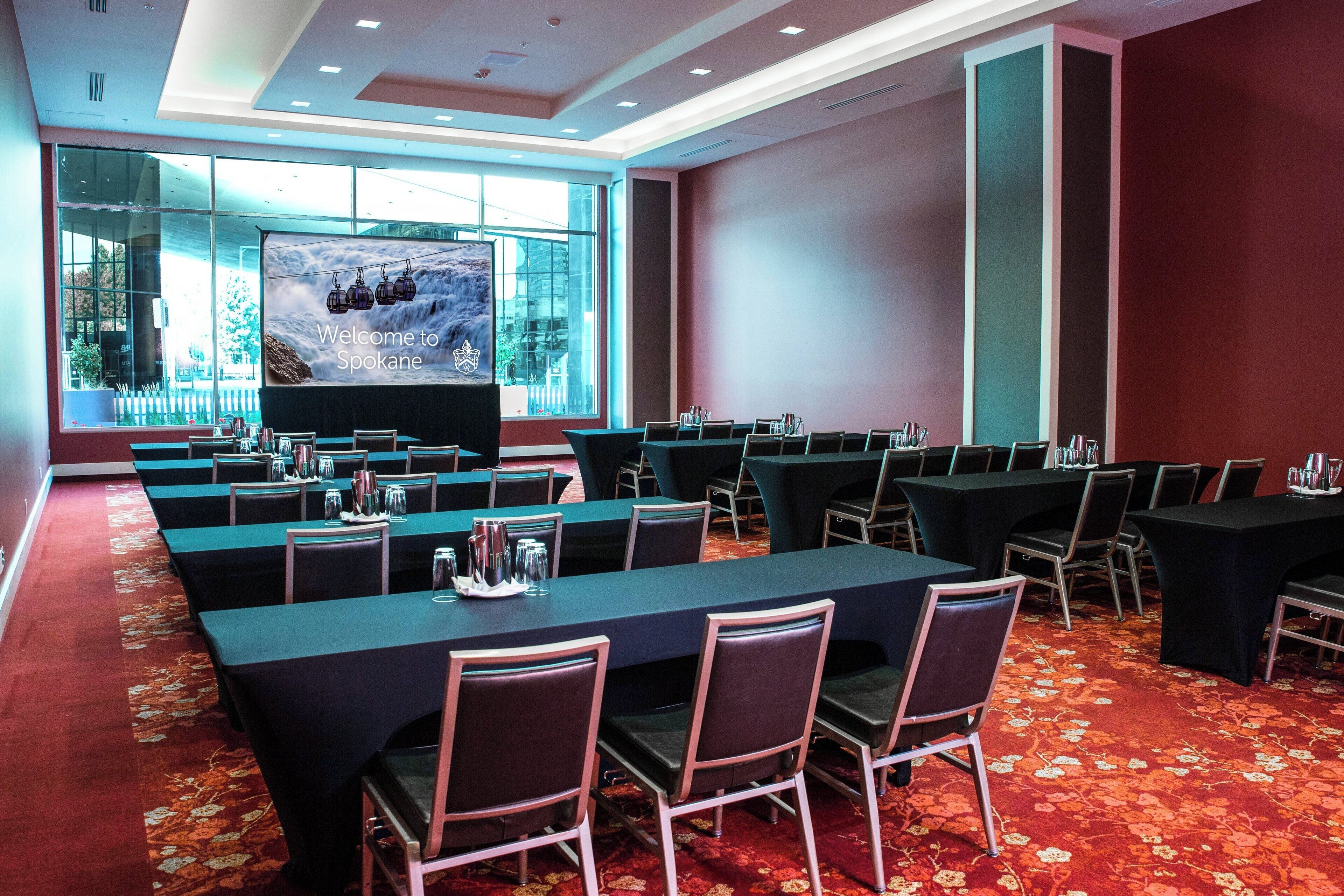 Une salle de conférence dotée de longues tables et de chaises, d’un grand écran affichant un message de bienvenue, et offrant une vue sur l’extérieur grâce à de grandes fenêtres.