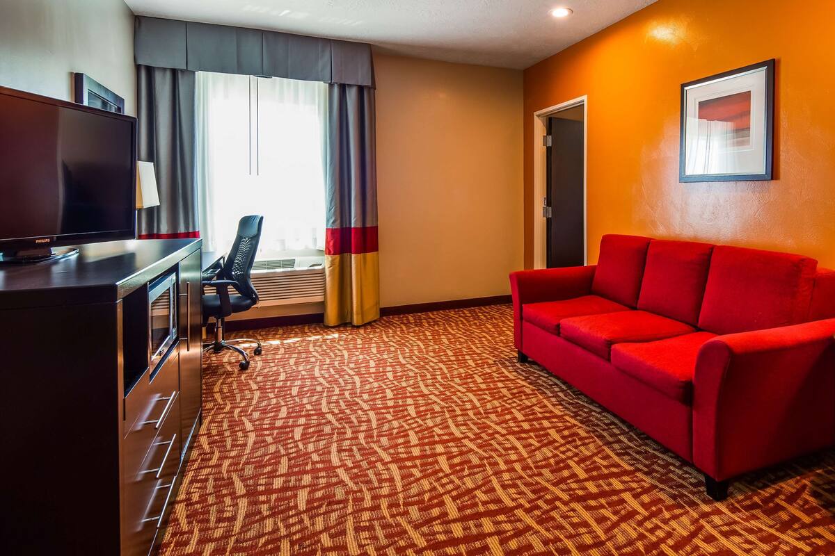 A hotel room with a red sofa, a television, a desk with a lamp, and a patterned carpet.