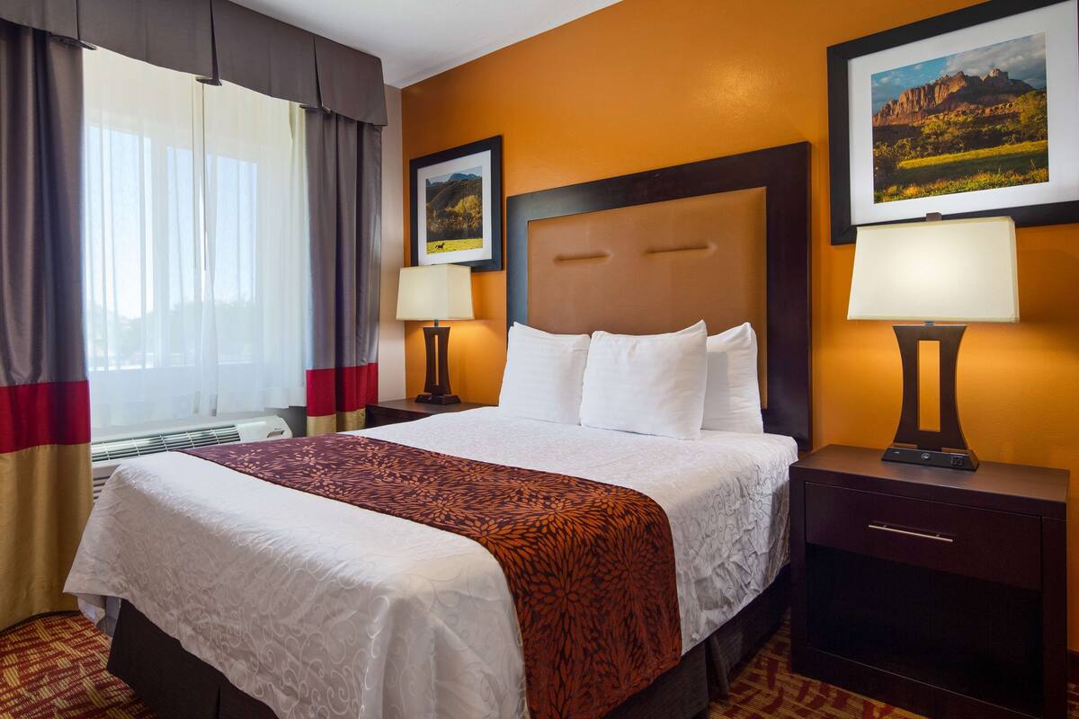 A hotel room with a large bed, two bedside tables with lamps, a window with curtains, and two framed pictures on the wall.