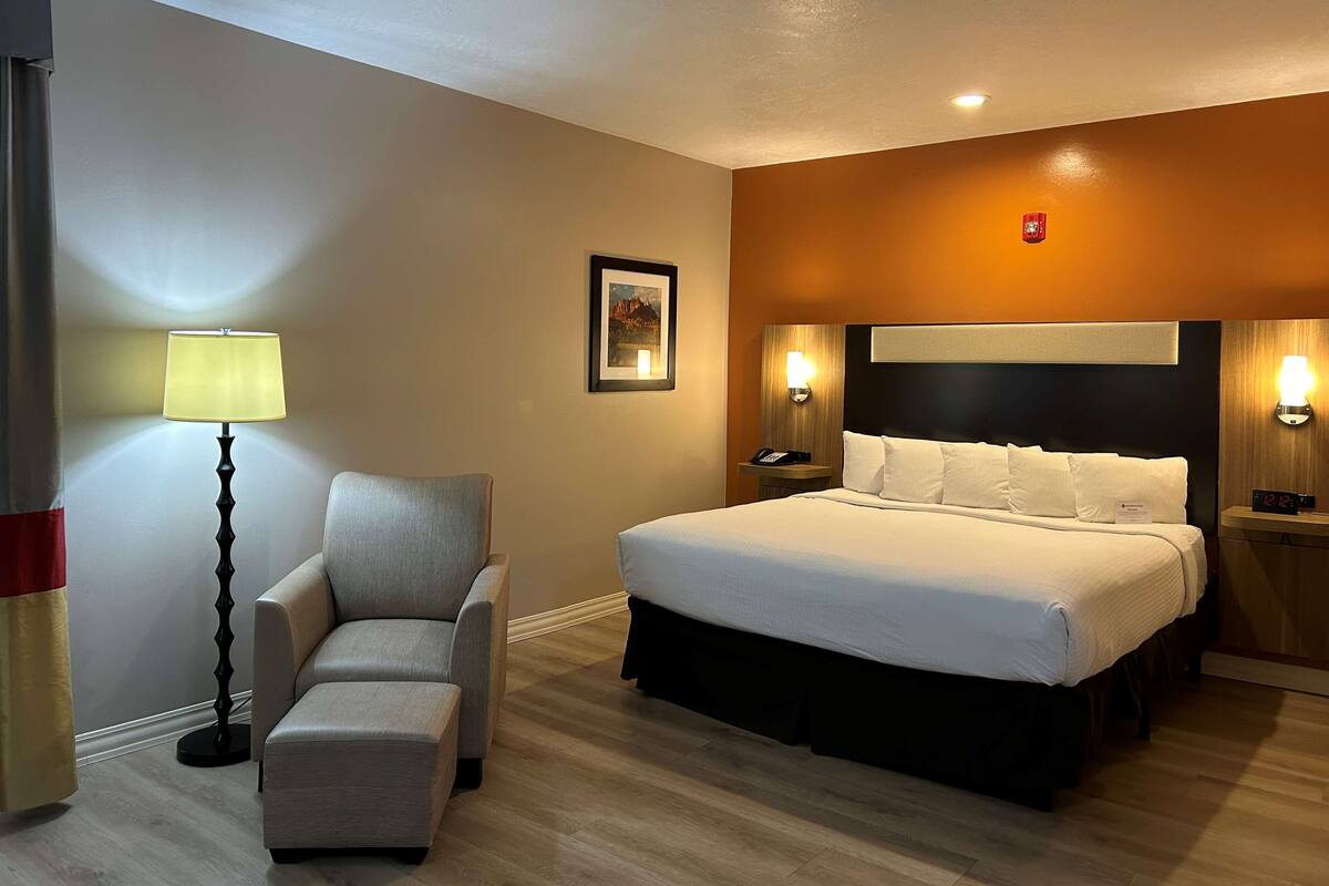 A hotel room with a bed, a chair, a bedside table, a lamp, and a wall-mounted clock.