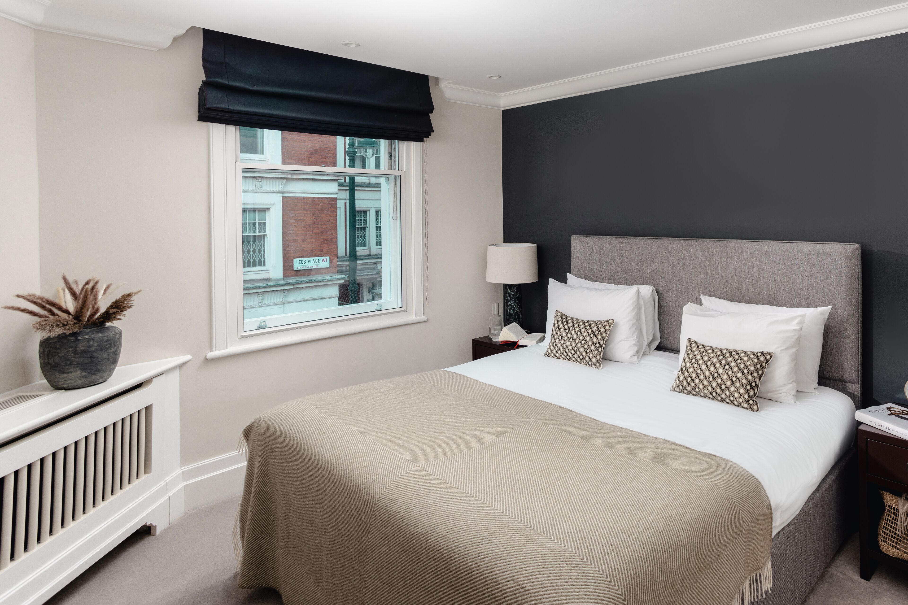 A neatly arranged bedroom with a large bed, a window with a view of buildings, a radiator, and a bedside table with a lamp and book.