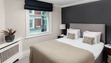 A neatly arranged bedroom with a large bed, a window with a view of buildings, a radiator, and a bedside table with a lamp and book.