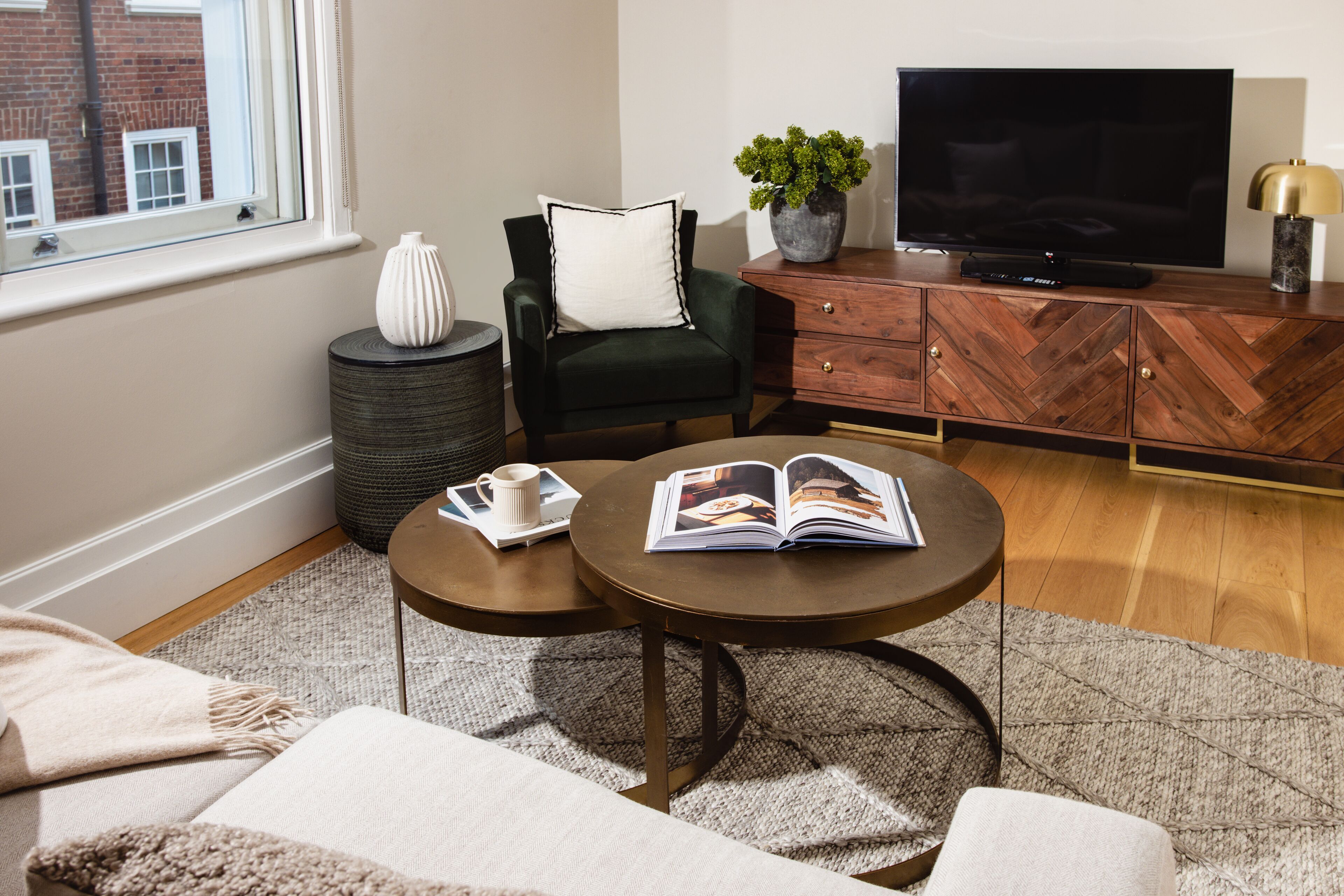 A living room with a sofa, two coffee tables, a television, a cabinet, and a window with a view of a brick building.