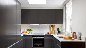 A modern kitchen with dark cabinets, a built-in oven, a countertop with a bread loaf, and a window with blinds. - Native Mayfair (London)