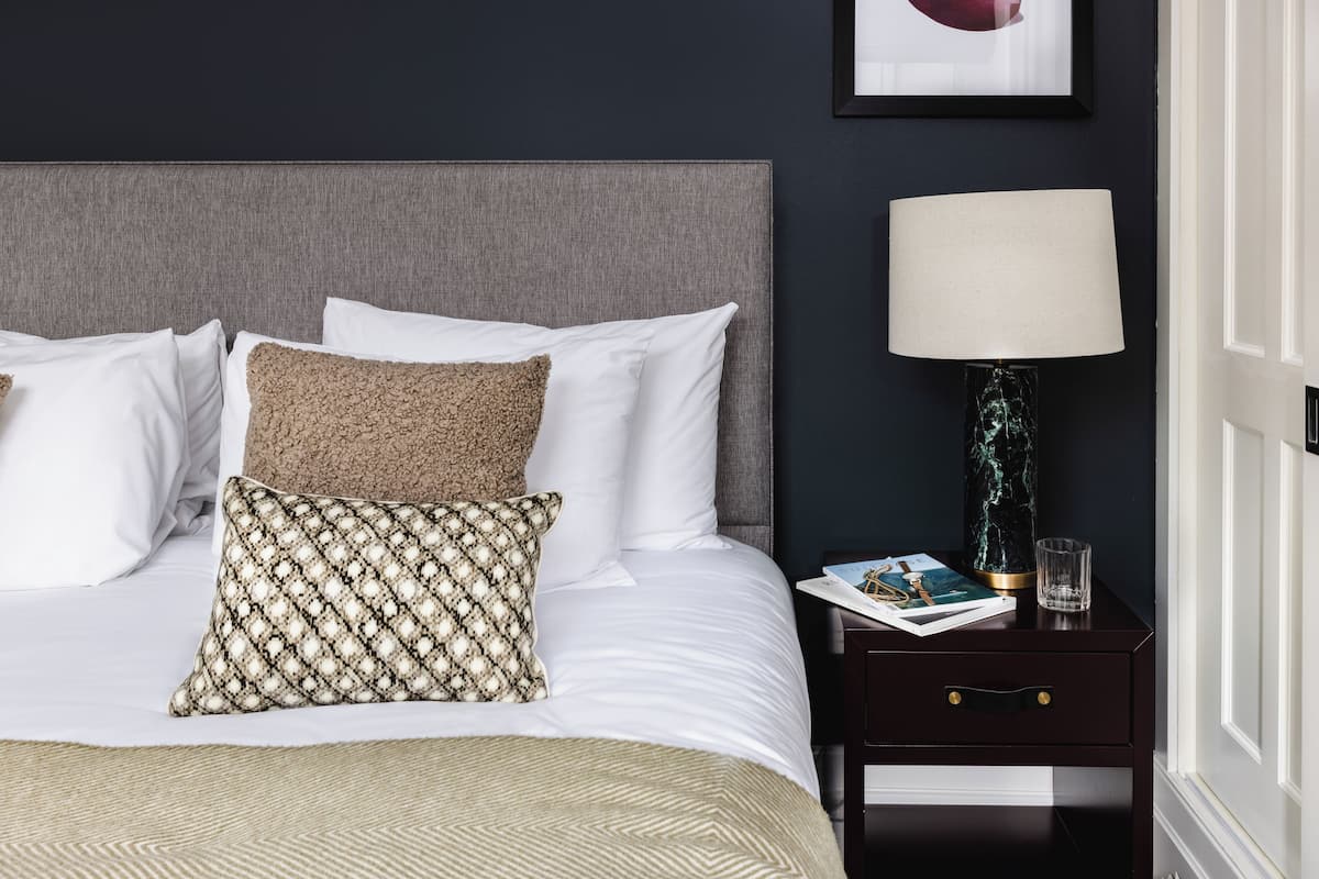 Una cama bien hecha con almohadas blancas y estampadas, una mesita de noche oscura con una lámpara y un cuadro en la pared.