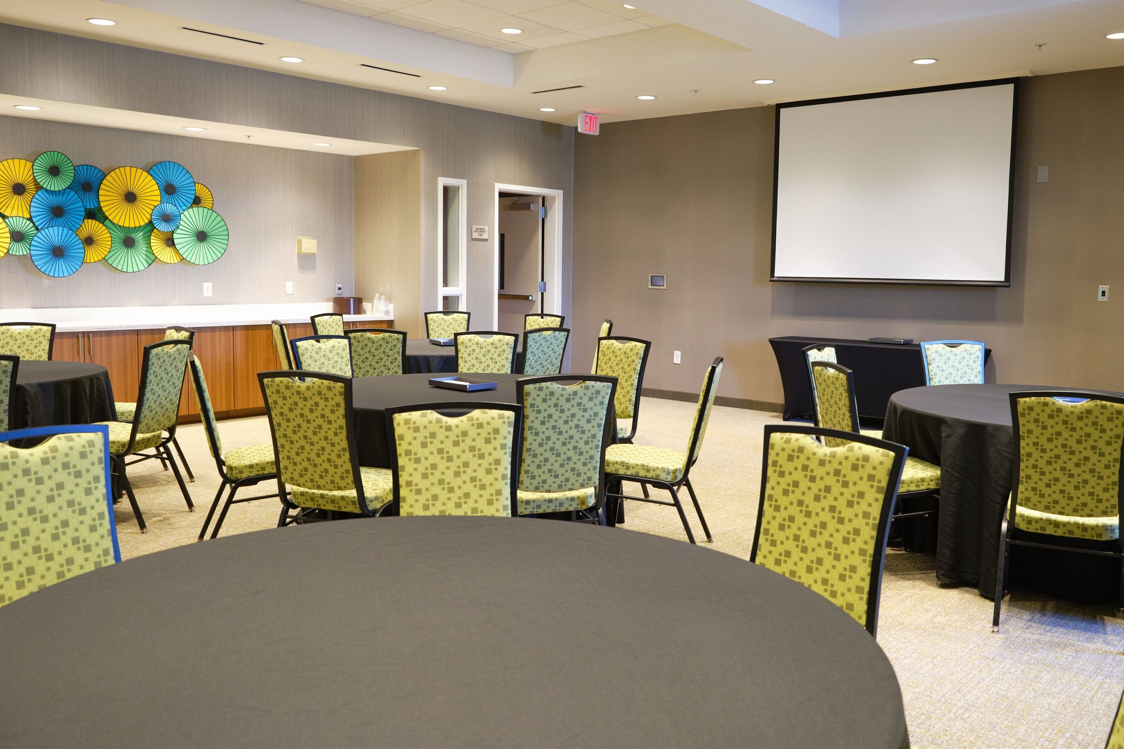 A conference room with a large table and chairs, a projector screen, and a wall with colorful umbrellas.